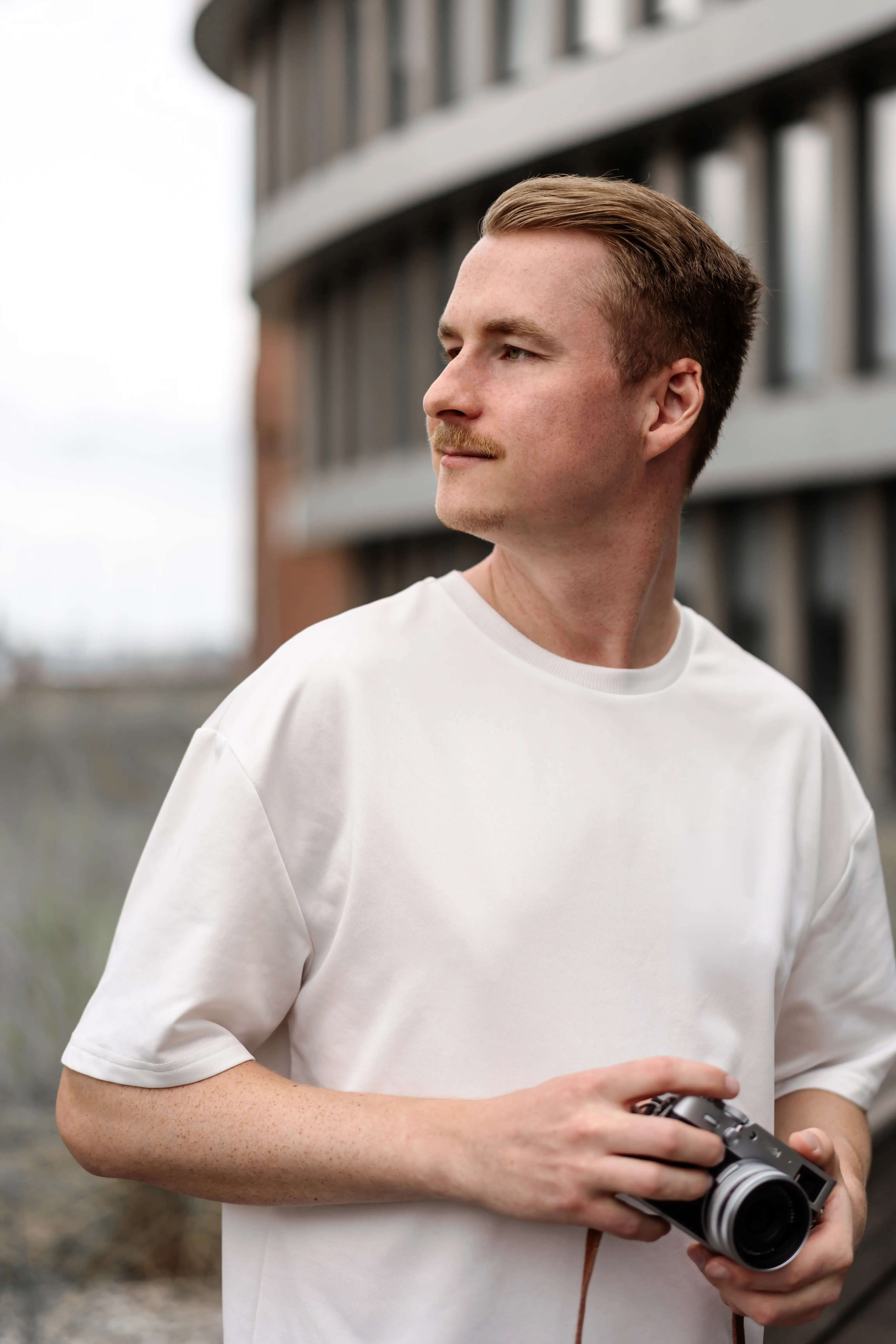 Ein professioneller Fotograf und Imagevideograf mit einer Kamera in der Hand, der vor einer modernen Unternehmensfassade in die Ferne blickt. Er trägt ein schlichtes weißes T-Shirt und steht für hochwertige Imagefotos und Imagevideos bereit. Ein professioneller Fotograf und Imagevideograf mit einer Kamera in der Hand, der vor einer modernen Unternehmensfassade in die Ferne blickt. Er trägt ein schlichtes weißes T-Shirt und steht für hochwertige Imagefotos und Imagevideos bereit.